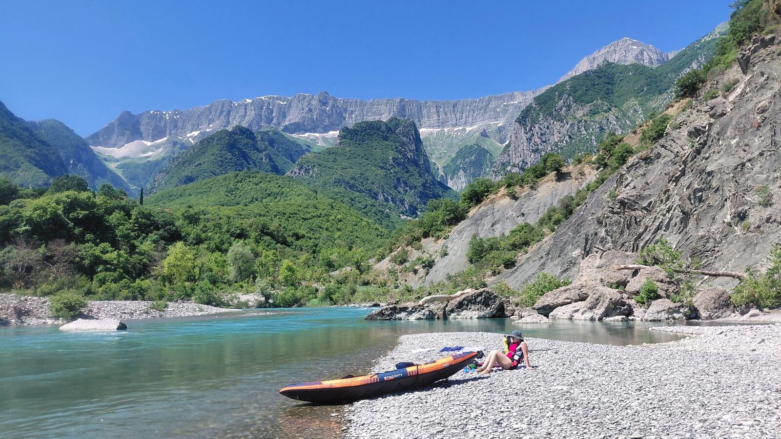 A kayak resting on a gravel riverbank beneath dramatic mountains in Albania